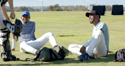 Two baseball teammates laughing and resting on grass during sunny outdoor practice session