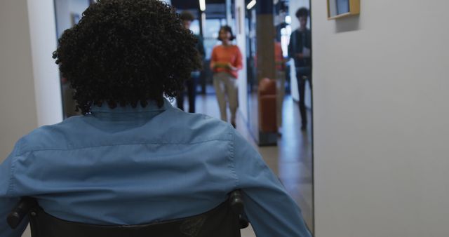 This image shows a person proceeding down a hallway in an office environment. The person's back is facing the camera as they navigate the space in a wheelchair. This photo can be used to highlight themes such as workplace diversity, inclusion, and accessibility. It is suitable for use in articles and materials that discuss workplace inclusivity, disability rights, and office design for better accessibility.