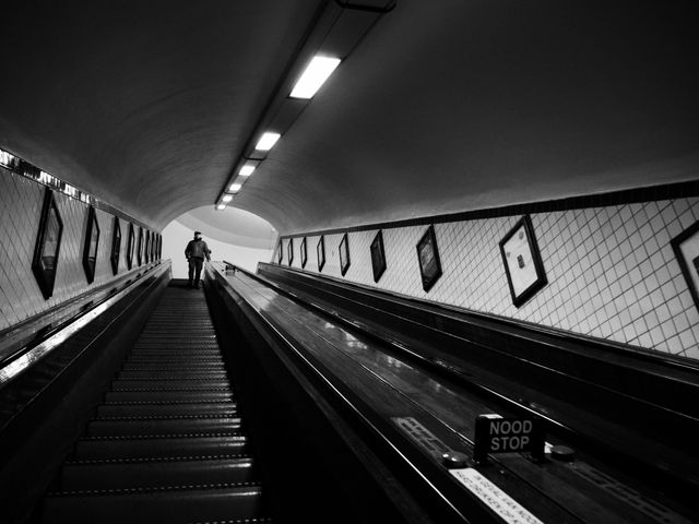 Person standing at the end of an escalator tunnel, viewed from bottom looking up, shot in black and white. This photo can be used in projects involving public transportation, urban life, and architectural design. Great for illustrating concepts of perspective, movement, and solitude.