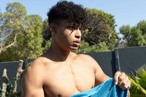 African american man standing shirtless holding blue towel and gazing in sunny backyard