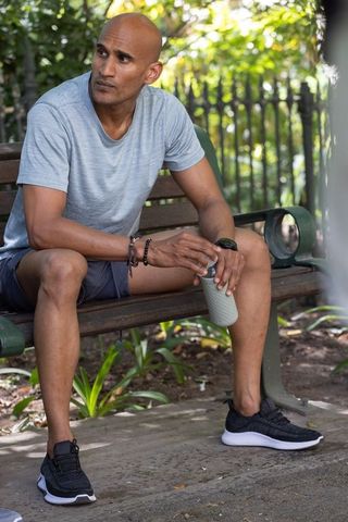 Fit african man sitting on park bench holding tumbler after outdoor run in athletic shorts