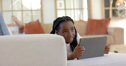 Happy african american girl lying on sofa using tablet for learning and play in bright living room