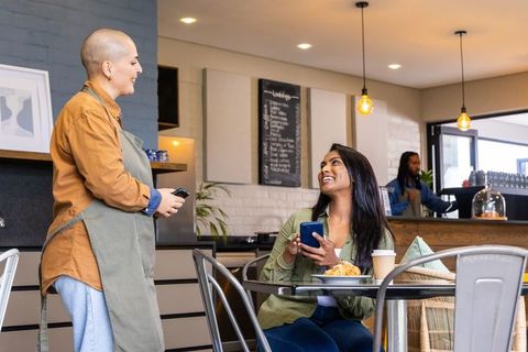 Indian woman enjoying coffee and croissant while chatting with friendly barista in urban cafe