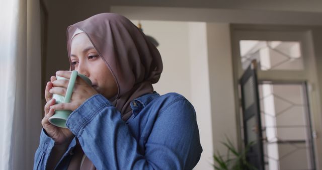 Image of happy biracial woman in hijab drinking coffee, looking out of window at home. Happiness, relaxation, inclusivity and domestic life.