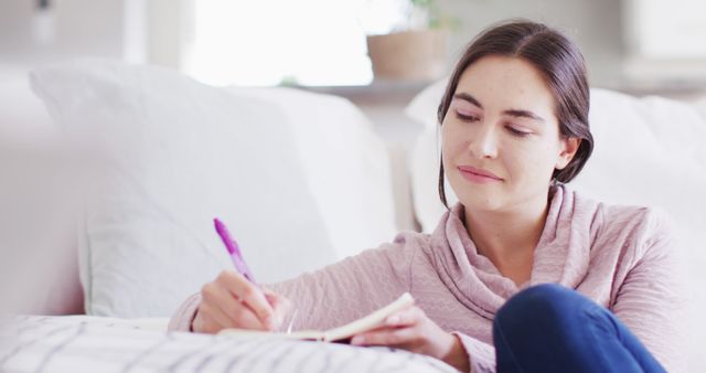 Young woman sitting on a couch, writing in notebook in a cozy home environment. Perfect for themes revolving around creativity, journaling, self-reflection, personal thoughts, or relaxation. Can be used in articles about home lifestyle, personal growth, or creative processes.