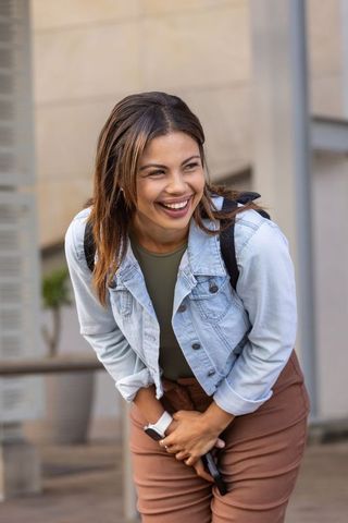 Laughing african woman holding smartphone at waist wearing denim jacket and backpack