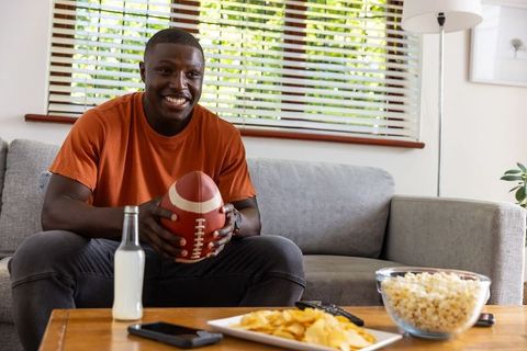 African american man smiling holding football on couch watching game with popcorn, chips