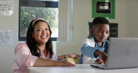 Female teacher mentoring African American boy using laptop in bright classroom