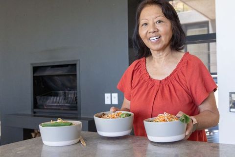 Senior asian woman serving three ceramic bowls of healthy salad in modern kitchen