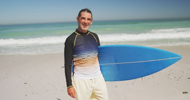 A man is holding a blue surfboard while standing on a sandy beach with ocean waves in the background. He is smiling and wearing a wetsuit, indicating that he is ready for a day of surfing. Suitable for use in marketing materials, travel advertisements, adventure sports promotions, and beach lifestyle content.