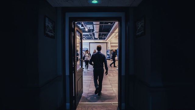 Man walking into bright lobby from dark corridor through open double doors, back silhouette