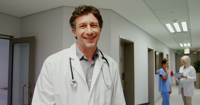 Doctor smiling confidently in a hospital hallway while medical staff discuss patient charts in background. Ideal for representing healthcare professional integrity, teamwork in a hospital environment, or medical facility services.