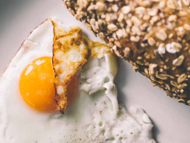 A sunny side up egg with a crispy edge sits next to a slice of whole grain bread with oats. The vibrant orange yolk and textured crust of the bread suggest a nutritious start to the day. Perfect for promoting healthy breakfast options, nutritious eating, or menu designs for cafes and restaurants.