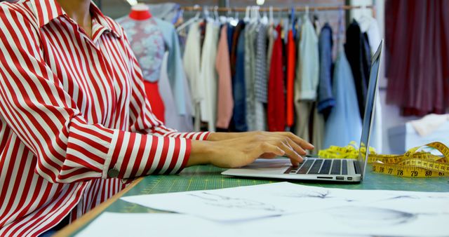 A fashion designer, middle-aged, is working on a laptop in a studio surrounded by garments, with copy space. Their focus on the screen suggests they are conceptualizing designs or managing business aspects of their fashion line.