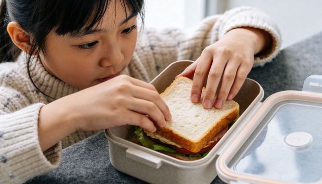 Young girl placing homemade sandwich into reusable lunchbox with clear orange-rim lid