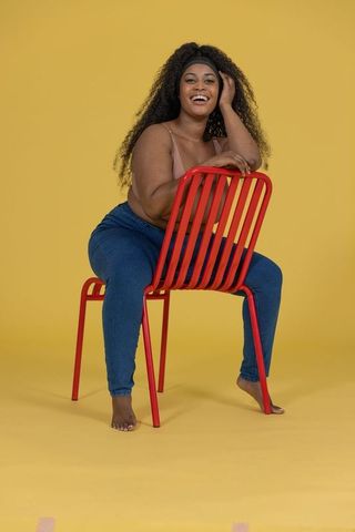Confident african-american woman leaning on red chair smiling on vibrant yellow backdrop