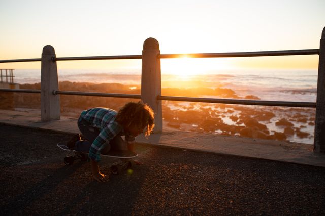 Boy kneeling on a skateboard on a promenade by the sea during sunset. Ideal for themes of childhood, outdoor activities, leisure, and adventure. Perfect for use in advertisements, blogs, and articles related to family fun, summer activities, and seaside vacations.