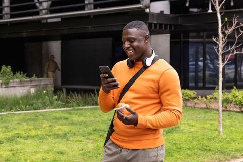African american man wearing orange sweatshirt and headphones checking phone and eating muffin