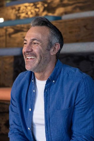 Middle-aged man laughing in cozy rustic lounge wearing blue shirt and warm smile