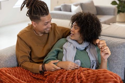 Couple cuddling on couch wrapped in rust-orange blanket sharing cozy conversation