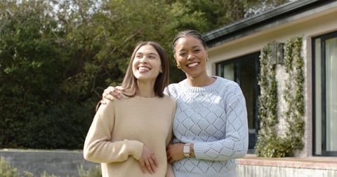 Smiling diverse women embracing on sunny lawn outside modern home wearing knit sweaters