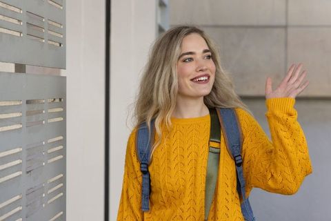 Young woman smiling and waving in mustard sweater with backpack against modern facade