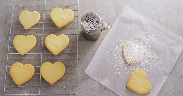 Heart-shaped cookies cool on a wire rack, with a sifter dusting one with powdered sugar, with copy space. Capturing a moment of baking, the image evokes feelings of love and the joy of homemade treats.