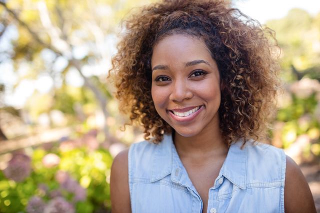 Young woman with curly hair smiling in a park. Ideal for use in lifestyle blogs, advertisements, or social media posts promoting outdoor activities, happiness, and natural beauty.