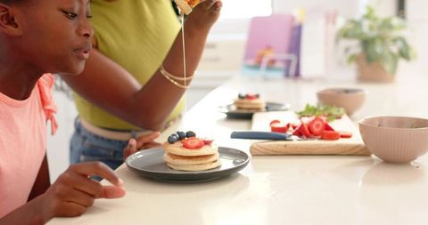 Mother and teen daughter pouring syrup on pancake stack with berries in bright kitchen