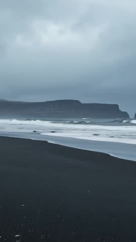Rolling waves lapping volcanic black sand beach with distant basalt cliff - vertical video