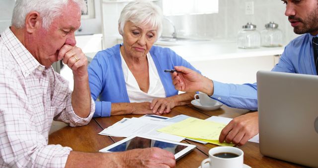 Senior couple sitting at table in home interior, meeting financial advisor to discuss investments and retirement planning. Advisor pointing at paperwork and digital tablet. Suitable for topics related to financial planning, retirement, senior advice, and professional consultations.
