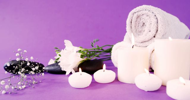 A serene spa setup features lit candles, fluffy towels, and smooth stones on a purple background, with copy space. Elements in the composition suggest relaxation and provide a visual representation of a tranquil wellness environment.