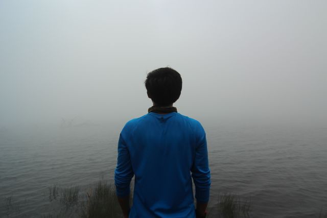 Man in a blue shirt standing at the edge of a foggy lake, facing the water. The mist and fog create a serene and tranquil environment. This image can be used for concepts related to solitude, reflection, serenity, and nature. Perfect for inspirational and motivational content, travel blogs, or mental health articles.