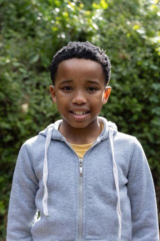 African american boy standing in gray hoodie and yellow tee with sunlit green foliage