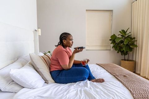 African american woman relaxing on bed sipping tea using remote in minimalist bedroom