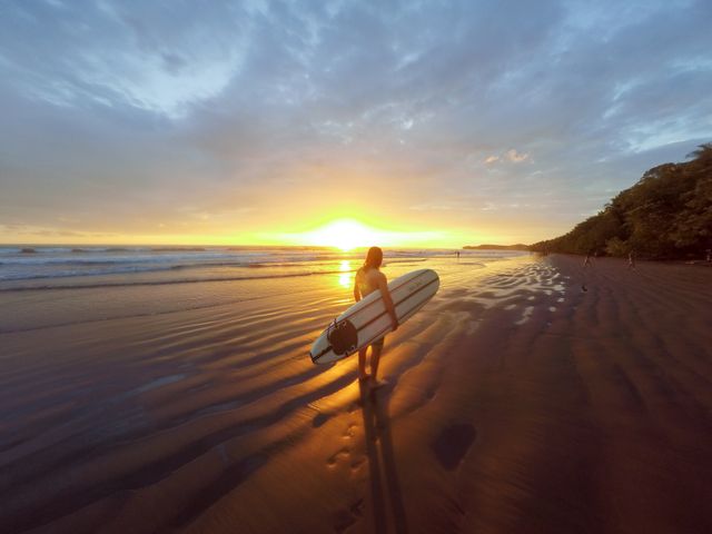 Silhouette of a surfer walking along the beach holding a surfboard at sunset, creating a tranquil and picturesque scene. Perfect for promoting coastal destinations, travel agencies, surf camps, and outdoor adventure brands. It evokes a sense of freedom, tranquility, and connection to nature.