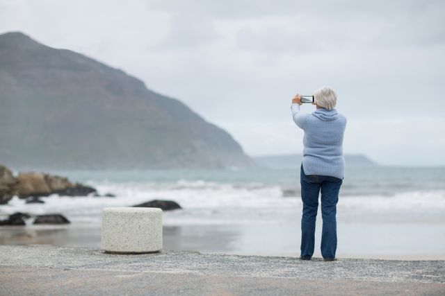 Senior woman standing on beach, capturing scenic ocean view with smartphone. Ideal for themes related to travel, retirement, leisure, technology use among seniors, and nature photography. Can be used in articles, blogs, and advertisements focusing on senior lifestyle, travel destinations, and outdoor activities.