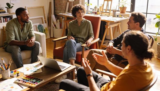 Creative team discussing art project in sunlit loft studio with laptop and palettes