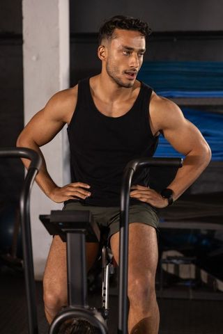 Muscular man pedaling air bike in gym wearing black tank top, green shorts and smartwatch