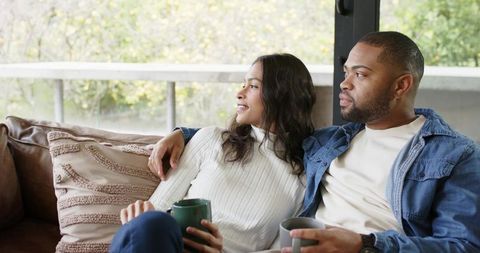 Diverse couple sitting on leather sofa holding mugs, cozy intimate morning by window
