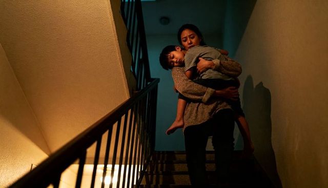 Asian mother carrying sleeping child down dim apartment stairwell, late-night caregiving