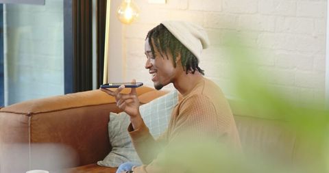 African American man speaking into smartphone while relaxing on leather sofa at home
