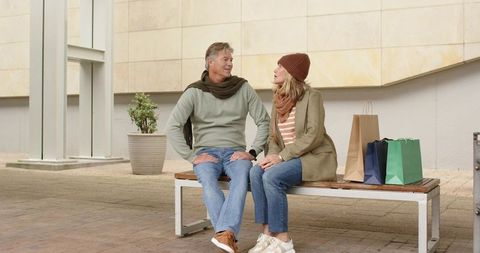 Mature couple sitting and chatting on bench after shopping in plaza wearing fall layers