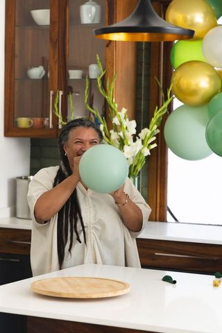 Mature woman inflating pastel green balloon in modern kitchen decorating for party