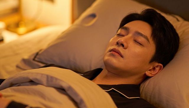 Sleeping man wearing dark pajamas under beige duvet on gray pillow in ambient bedroom