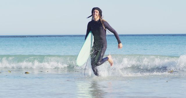 Female surfer running with surfboard in hand at ocean beach. Great for advertisements focused on water sports, active lifestyle promotions, and travel destinations targeting surfing enthusiasts. Ideal for use in websites and brochures encouraging outdoor and adventure activities.