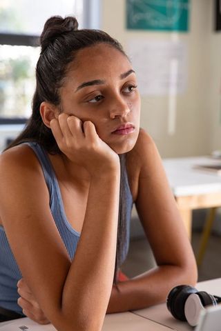 Pensive teenage female student leaning on desk, wearing blue top, headphones nearby