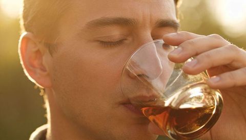 Man sipping whiskey from clear tumbler during golden hour, glass reflecting warm rimlight