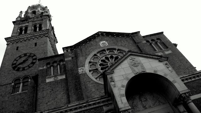 This image showcases a historic church building with a prominent clock tower, captured in a black and white modern photograph. The church's architecture exhibits Gothic and Romanesque elements with intricate stone carvings and a large clock on the tower. Use this image for storytelling content involving historical architecture, religious history, photography art, and educational materials about architectural styles and heritage.