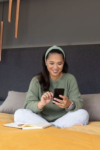 Mid-adult asian woman sitting on bed smiling and tapping smartphone in cozy bedroom
